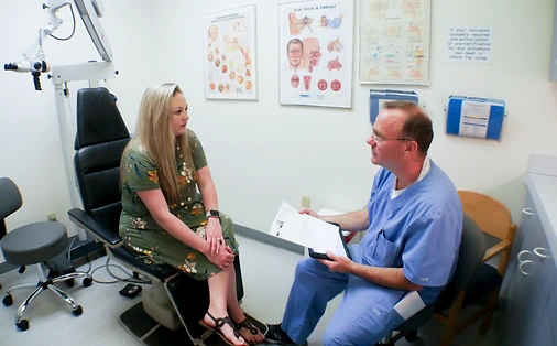 ENT specialist in blue scrubs consulting with a patient in an examination room with anatomical diagrams on the wall
